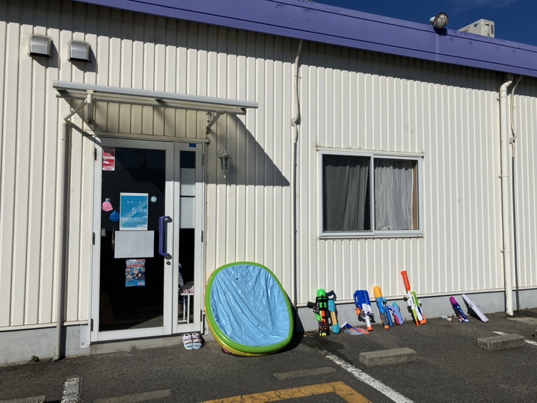 白い波板の壁にある入口。ひさしの下のガラス戸に「5」と書かれた紙がはられ、手前にビニールプールと水鉄砲がならんでいる