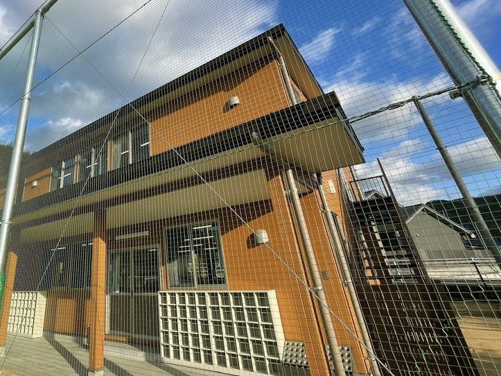 オレンジ色のタイルの2階建ての建物。1階に大きなガラス戸と下駄箱がならび、手前に打球よけの高いネットが張られている