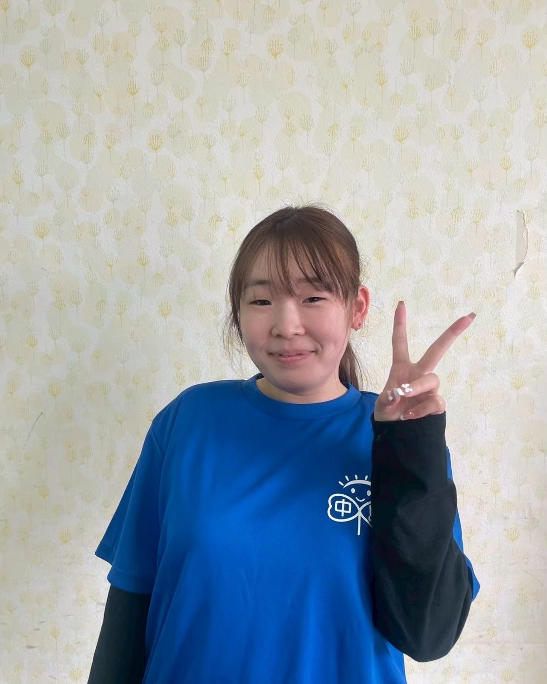花柄の壁を背に、片手でピースをしている支援員