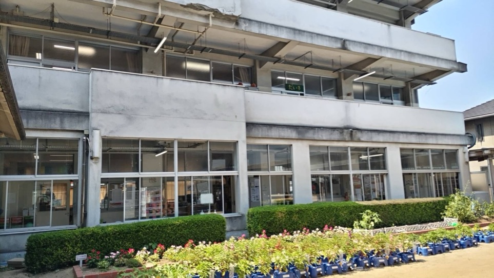 とみた児童クラブが部屋を借りている富田小学校の校舎。2階建ての白い建物で、1階と2階に大きな窓がならび、手前に生垣と花のプランターが置かれている